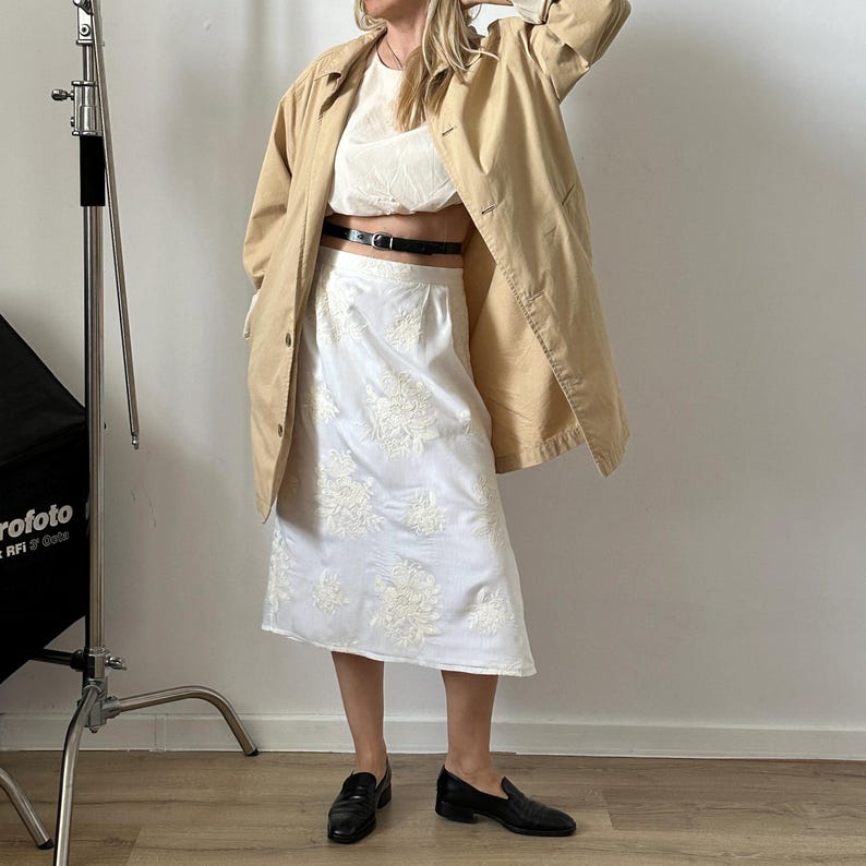 Person wearing a beige jacket, white top, and white skirt with floral embroidery indoors.