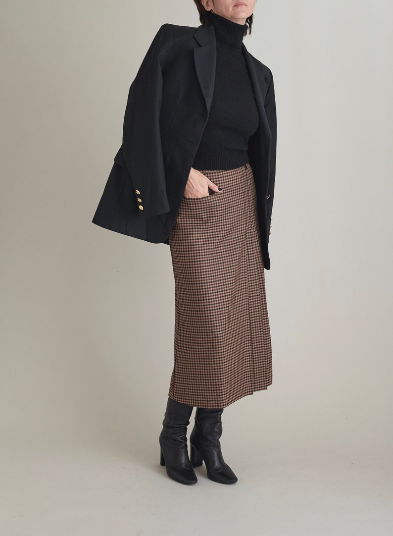 Person wearing a black coat, black turtleneck, brown checkered skirt, and black boots on a plain background