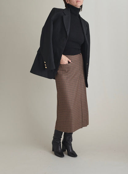 Person wearing a black coat, black turtleneck, brown checkered skirt, and black boots on a plain background