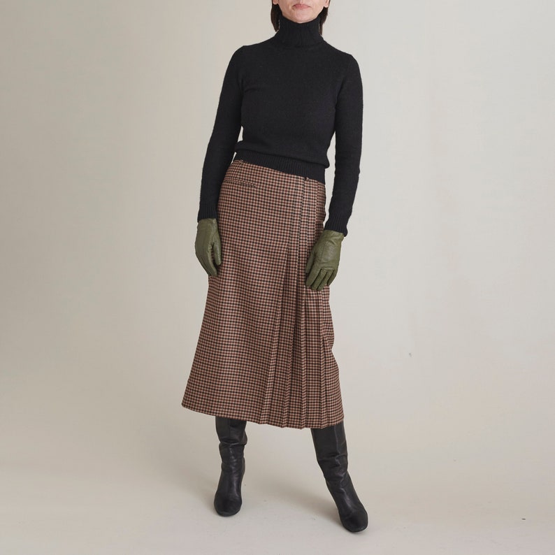 Person wearing a black turtleneck, brown plaid skirt, green gloves, and black boots on a beige background